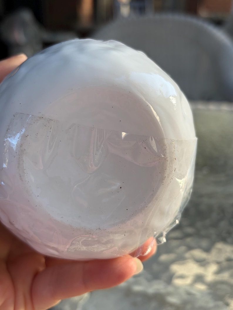 May include: A white, round ceramic object, possibly a bowl or cup, wrapped in clear plastic. The object is smooth and appears to be new, with a clean, pristine surface. The plastic wrap is secured with clear tape.