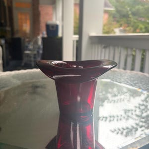 May include: A deep red glass vessel featuring a wide, flared, and undulating rim that tapers down to a narrower, circular base. The translucent material allows light to pass through, highlighting the rich crimson hue and smooth, polished surface.