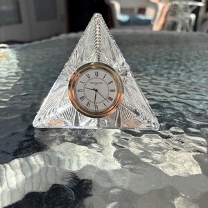 May include: A clear crystal triangle clock with a gold-tone clock face. The clock face has black Roman numerals and the word "Waterford" printed on it. The clock is sitting on a glass table.