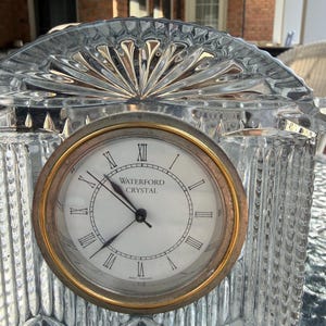 K&ouml;nnte beinhalten: Eine transparente Kristalluhr mit goldfarbener L&uuml;nette und wei&szlig;em Zifferblatt. Das Zifferblatt hat schwarze r&ouml;mische Ziffern und die Worte "WATERFORD CRYSTAL". Die Uhr hat eine dekorative, f&auml;cherf&ouml;rmige Oberseite.