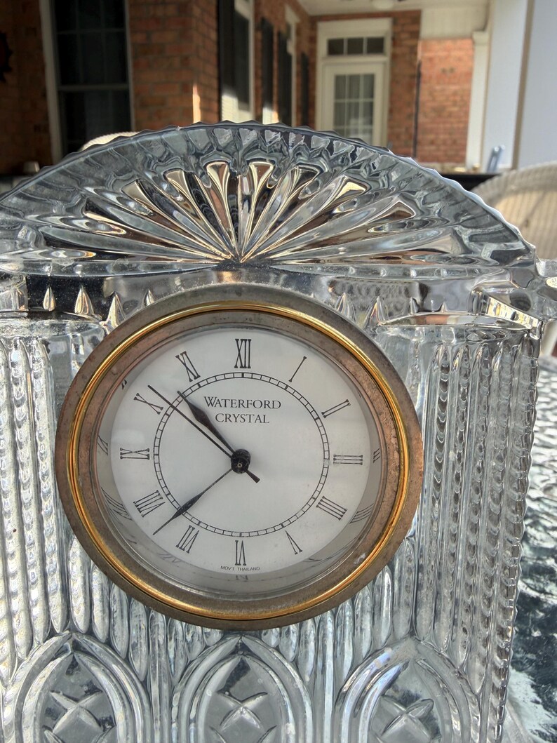 K&ouml;nnte beinhalten: Eine transparente Kristalluhr mit goldfarbener L&uuml;nette und wei&szlig;em Zifferblatt. Das Zifferblatt hat schwarze r&ouml;mische Ziffern und die Worte "WATERFORD CRYSTAL". Die Uhr hat eine dekorative, f&auml;cherf&ouml;rmige Oberseite.