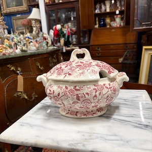 May include: A white ceramic tureen with a lid, featuring a red floral pattern. The tureen has handles and a heart-shaped opening on the lid. It sits on a marble-topped table, showcasing a vintage aesthetic.