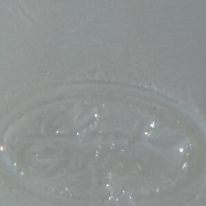 May include: Close-up of a clear, textured glass object with an oval shape. The glass has a slightly frosted appearance and reflects light, creating small, bright highlights. The object appears to be part of a larger glass item.