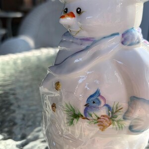 May include: A ceramic snowman figurine with a white body, a carrot nose, and a painted blue bird on its side. The snowman wears a hat and a scarf with blue and purple accents. The figurine has a glossy finish.