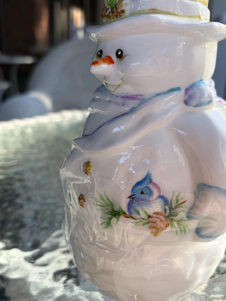 May include: A ceramic snowman figurine with a white body, a carrot nose, and a painted blue bird on its side. The snowman wears a hat and a scarf with blue and purple accents. The figurine has a glossy finish.