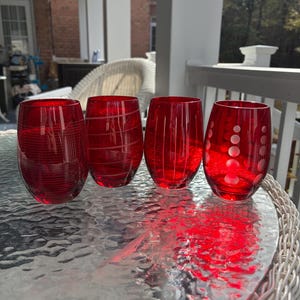 May include: Four red glass tumblers with varying etched designs. The tumblers are displayed on a glass table, with a wicker table edge visible. The etched patterns include vertical lines, swirls, and polka dots. The glasses are a vibrant red color.