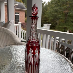 May include: A tall, decorative glass decanter with a red and clear design. The decanter features a red stopper and intricate patterns. The design includes vertical red stripes and a diamond-like pattern. The decanter is likely used for serving beverages.