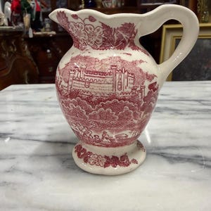 May include: A white ceramic pitcher with a red transferware design. The pitcher features a castle scene and floral patterns. It has a curved handle and a scalloped rim. The pitcher is approximately 8 inches tall.