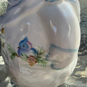 May include: White ceramic jar with a lid, decorated with a blue bird perched on a branch with green leaves and a brown pinecone. The jar has blue and purple accents and is wrapped in clear plastic.