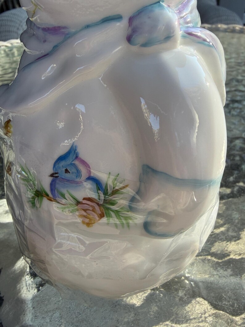 May include: White ceramic jar with a lid, decorated with a blue bird perched on a branch with green leaves and a brown pinecone. The jar has blue and purple accents and is wrapped in clear plastic.