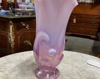 May include: A tall, antique, pink glass vase with a ruffled rim. The vase has a flared shape and a rounded base. Swirls and decorative elements adorn the sides. The vase is displayed on a marble-topped table.