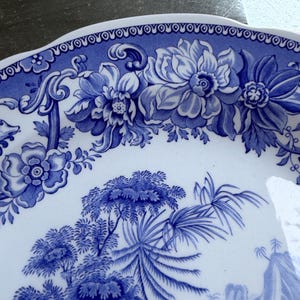 May include: A white ceramic plate featuring a blue transferware pattern. The rim displays a decorative border of stylized flowers and scrolling foliage, while the center depicts a detailed landscape with trees, mountains, and lush vegetation.