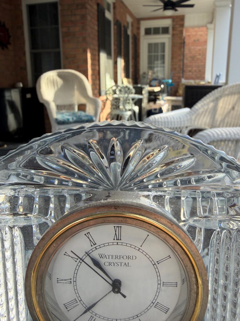 K&ouml;nnte beinhalten: Eine transparente Waterford Crystal-Uhr mit goldfarbener L&uuml;nette und r&ouml;mischen Ziffern. Das Zifferblatt zeigt "WATERFORD CRYSTAL". Die Uhr befindet sich vor einem verschwommenen Hintergrund im Freien.