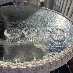 May include: Four small, clear glass dishes with a starburst design are arranged on a glass-topped table. A matching pitcher sits in the background. The table has a white wicker edge, and the scene is outdoors.