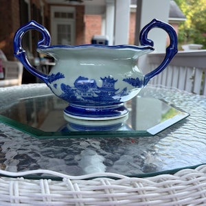 May include: A white ceramic sugar bowl featuring a blue willow pattern with pagodas and trees. The bowl has a scalloped rim, a circular base, and two prominent, curved blue handles extending from the sides, creating a classic two-handled design.