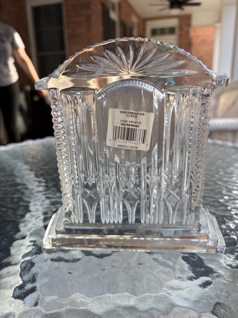 K&ouml;nnte beinhalten: Eine transparente Westminster-Uhr aus Bleikristall mit rechteckigem Sockel und dekorativer Oberseite. Die Uhr hat vertikale und geometrische Muster. Auf dem Etikett steht "Westminster Clock" und "Lead Crystal".