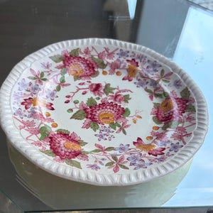 May include: A white ceramic plate with a floral design. The plate features a detailed pattern of pink, red, yellow, and blue flowers with green leaves. The rim has a textured, ridged design. The plate is likely vintage or antique.