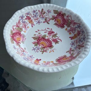 May include: White ceramic bowl with a scalloped rim, featuring a floral pattern in shades of pink, red, yellow, and blue. The bowl is set on a light green base, reflecting on a glass surface. A decorative piece for serving or display.