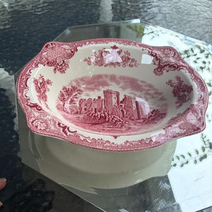 May include: An oval ceramic bowl featuring a deep red transferware pattern on a cream background. The center depicts a detailed landscape with a castle ruin, while the wide, scalloped rim is decorated with intricate floral and scroll motifs.