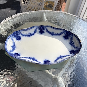May include: An oval, white ceramic serving platter with a scalloped edge. The platter features a blue floral design around the rim. The platter is sitting on a glass table.
