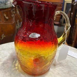 May include: A handcrafted glass pitcher with a crackle finish. The pitcher transitions from a deep red at the top to yellow at the base. It has a clear, curved handle and a spout for pouring. The label reads "BLENKO HANDCRAFT".