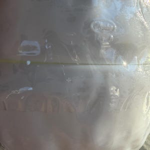 May include: A clear, round glass bottle with a textured surface. The bottle is wrapped in clear plastic. The bottle has a slight pearlescent sheen and is partially filled with a white liquid.