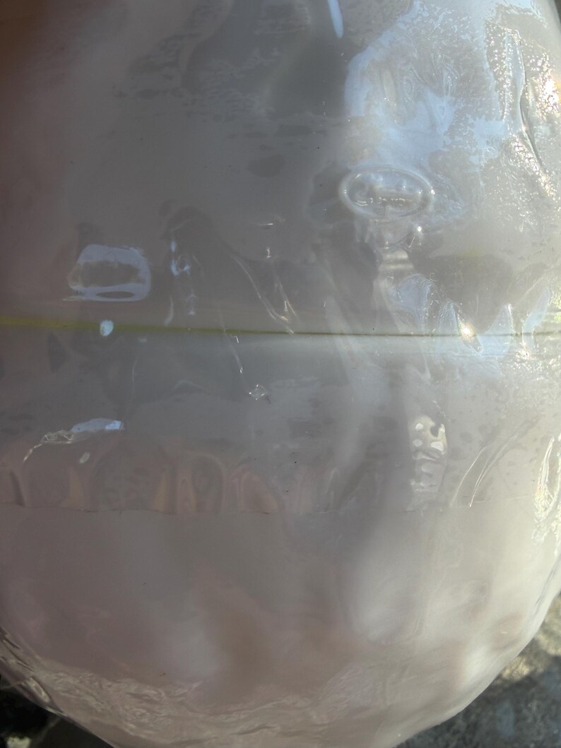 May include: A clear, round glass bottle with a textured surface. The bottle is wrapped in clear plastic. The bottle has a slight pearlescent sheen and is partially filled with a white liquid.