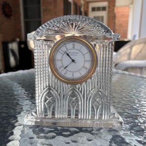 K&ouml;nnte beinhalten: Eine klare Kristalluhr mit goldfarbenem Rahmen und wei&szlig;em Zifferblatt. Die Uhr hat r&ouml;mische Ziffern und die Worte "WATERFORD CRYSTAL" auf dem Zifferblatt. Die Uhr ist rechteckig mit gew&ouml;lbter Oberseite.