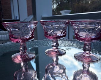 May include: Three pink vintage stemmed glasses with a decorative pattern. The glasses have a flared bowl, a twisted stem, and a round base. The glasses are sitting on a reflective surface, with their reflections visible below.