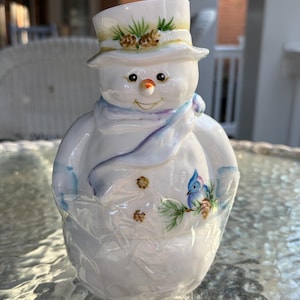 May include: A ceramic snowman figurine with a top hat adorned with pine needles and cones. The snowman has a carrot nose, a blue scarf, and a painted bluebird on its body. The figurine is white with colorful accents.