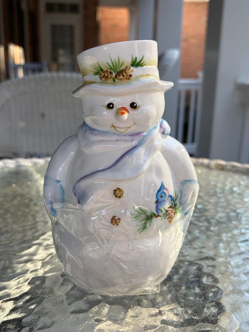 May include: A ceramic snowman figurine with a top hat adorned with pine needles and cones. The snowman has a carrot nose, a blue scarf, and a painted bluebird on its body. The figurine is white with colorful accents.