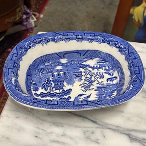 May include: Oval blue and white ceramic dish with a Willow pattern design. The dish features a detailed landscape scene with bridges, buildings, and trees. The rim has a decorative border. The dish is placed on a marble surface.