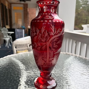 May include: A ruby red glass vase with etched botanical designs. The vase features a flared top with a series of small, circular cutouts. The body of the vase is adorned with etched trees and foliage. The vase sits on a round, faceted base.