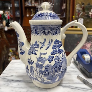 May include: A white ceramic teapot with a blue Willow pattern design. The teapot features a lid, spout, and handle. The blue pattern includes pagodas, trees, and birds. The teapot is sealed in plastic.