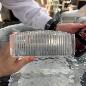 K&ouml;nnte beinhalten: Ein klarer, rechteckiger Glasaschenbecher mit vertikalen Rippendetails. Der Aschenbecher wird von einer Person gehalten, mit einem verschwommenen Hintergrund einer Au&szlig;enumgebung.