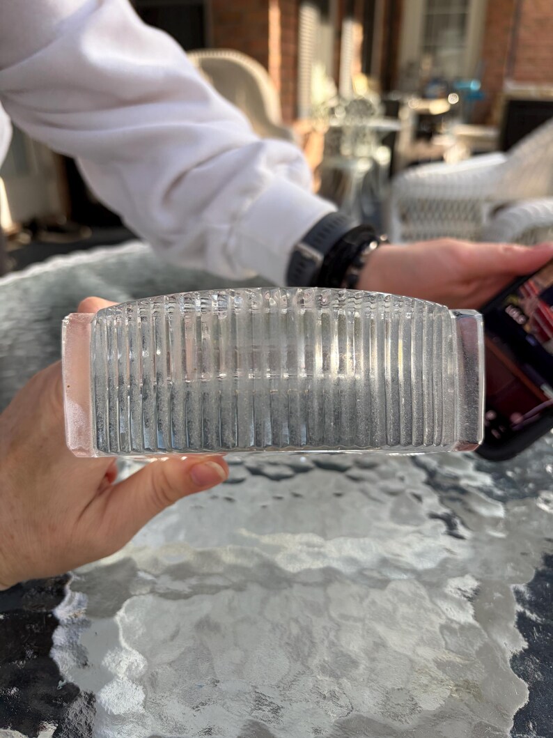 K&ouml;nnte beinhalten: Ein klarer, rechteckiger Glasaschenbecher mit vertikalen Rippendetails. Der Aschenbecher wird von einer Person gehalten, mit einem verschwommenen Hintergrund einer Au&szlig;enumgebung.