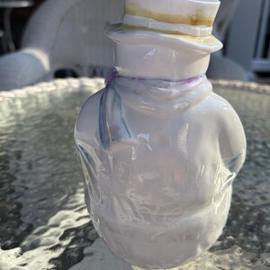 May include: A white ceramic snowman figurine wearing a top hat with a yellow brim and a light blue scarf. The snowman has a glossy finish and is standing on a glass table.