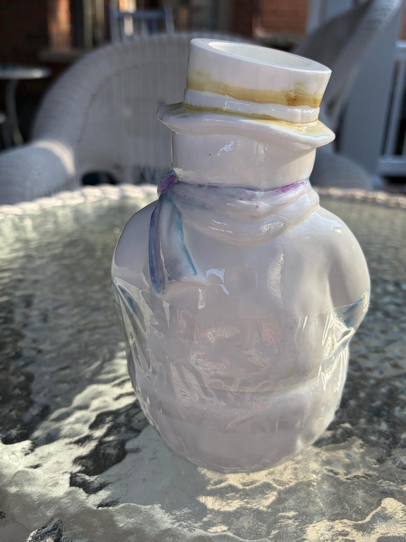 May include: A white ceramic snowman figurine wearing a top hat with a yellow brim and a light blue scarf. The snowman has a glossy finish and is standing on a glass table.