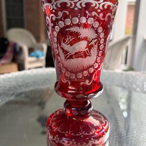 May include: A ruby red glass goblet featuring intricate white etched detailing. The bowl displays a central scene of a leaping deer surrounded by a circular border of dots and scrolling patterns. The base also features etched decorative motifs.