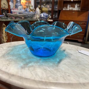 May include: A vibrant, turquoise-blue glass bowl with a ruffled rim. The bowl is round and sits on a marble-topped table. The glass is transparent, allowing light to pass through, creating a play of shadows and reflections.