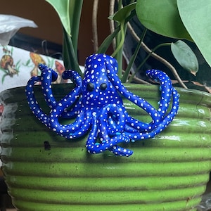 May include: A blue and white polka dot octopus figurine sits on a green ceramic pot with a wooden plant stand. The pot is on a wooden shelf with a green plastic corner brace.