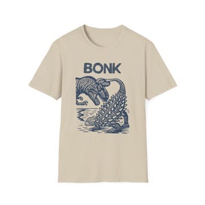 Pode incluir: Camiseta bege com um gráfico azul de um T-Rex e um Anquilossauro, com a palavra "BONK" acima. Os dinossauros são ilustrados em estilo cartoon, com o Anquilossauro voltado para o T-Rex, em um fundo aquático.