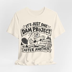 Puede incluir: Una camiseta de manga corta color crema con un gráfico negro de un castor cargando un tronco junto a una pila de leña. El texto "IT'S JUST ONE DAM PROJECT AFTER ANOTHER" aparece en pancartas sobre y debajo de la ilustración central.
