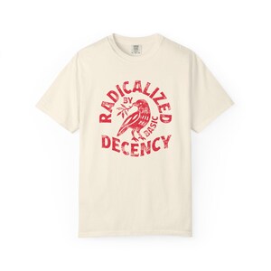 Könnte beinhalten: Ein cremefarbenes Kurzarm-T-Shirt mit einer roten kreisförmigen Grafik auf der Brust. Das Design zeigt einen Vogel, der einen Zweig im Schnabel hält, umgeben von dem Text "RADICALIZED BY BASIC DECENCY" in einer abgenutzten, verwitterten Schriftart.