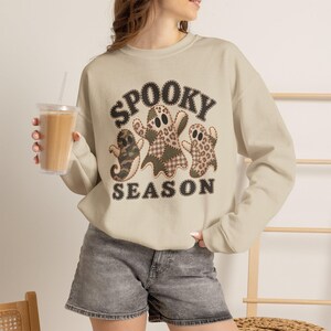 May include: A beige crewneck sweatshirt featuring a graphic of three ghosts with various brown and tan patterns, including leopard print and plaid. The text "SPOOKY SEASON" is printed in a dark brown, stitched-style font above and below the ghosts.
