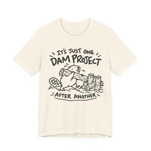 May include: A cream-coloured short-sleeve t-shirt featuring a black line-art graphic of a beaver carrying a log next to a pile of wood. The text above and below the beaver reads, IT'S JUST ONE DAM PROJECT AFTER ANOTHER, in a bold, hand-drawn style font.