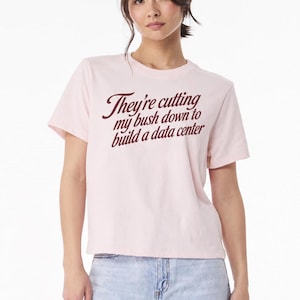 Könnte beinhalten: Ein hellrosa T-Shirt mit kurzen Ärmeln und dem dunkelroten Text "They're cutting my bush down to build a data center" auf der Brust. Das Kleidungsstück hat einen Rundhalsausschnitt und einen geraden Saum, kombiniert mit einer hellblauen Jeans.