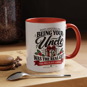 May include: A white ceramic mug with a red interior and handle, featuring black and red text that reads: MERRY CHRISTMAS 2026 BEING YOUR UNCLE WAS THE REAL GIFT THIS MUG IS JUST A BONUS HOLIDAY EDITION, decorated with a gift box and holly.