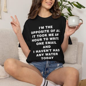 Könnte beinhalten: Ein schwarzes T-Shirt mit kurzen Ärmeln und weißem Text: I'M THE OPPOSITE OF AI, IT TOOK ME AN HOUR TO WRITE ONE EMAIL AND I HAVEN'T HAD ANY WATER TODAY. Das Shirt hat einen Rundhalsausschnitt und eine lockere Passform aus einfarbigem schwarzem Stoff.