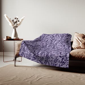 Puede incluir: Una manta suave con un fondo de color lavanda claro y un patrón botánico repetitivo de color morado oscuro con diversas hojas, tallos y pequeñas flores, colocada sobre el borde de un sofá de cuero marrón en una sala de estar.